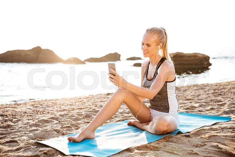 Attractive happy young woman doing yoga and using cell phone sitting on mat outdoors on beach, stock photo