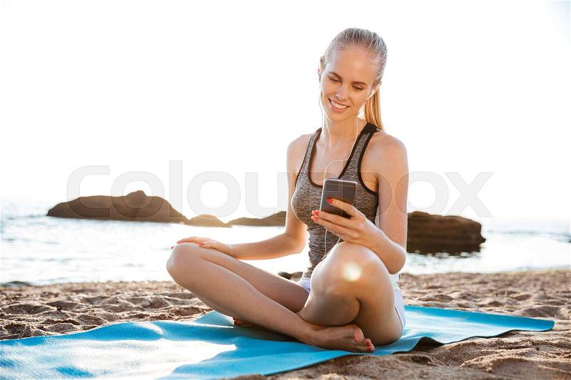 Attractive happy young woman doing yoga and using cell phone sitting on mat outdoors on beach, stock photo