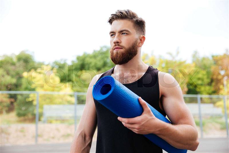 Concentrated serious male yoga instructor holding mat while standing outdoors, stock photo