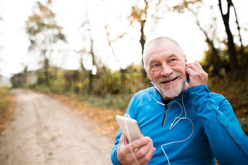 Senior runner in nature. Man with smart phone with earphones. Listening music or using a fitness app. Using phone app for tracking weight loss progress, running goal or summary of his run, stock photo