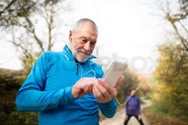 Senior runners in nature, stretching. Man with smart phone with earphones. Listening music or using a fitness app. Using phone app for tracking weight loss progress, running goal or summary of his run, stock photo