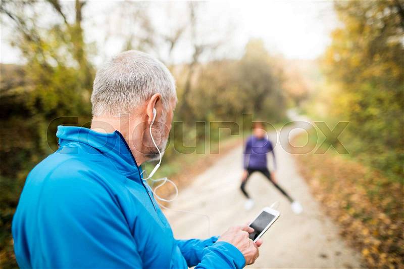 Senior runners in nature, stretching. Man with smart phone with earphones. Listening music or using a fitness app. Using phone app for tracking weight loss progress, running goal or summary of his run, stock photo