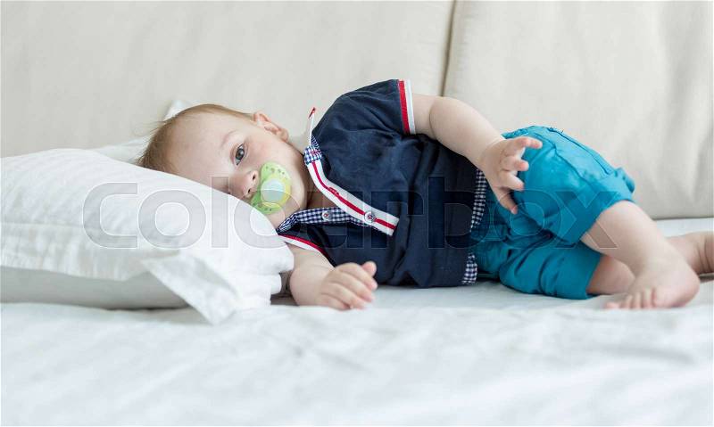 Cute baby boy with soother lying on big pillow on sofa, stock photo