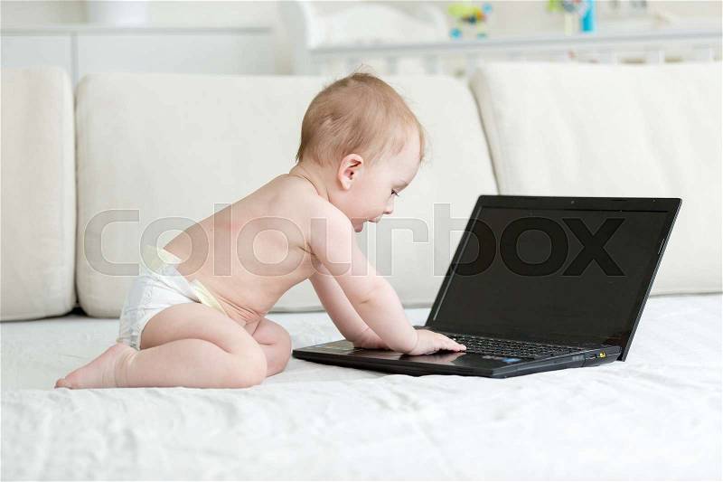 Adorable 1 year old baby boy sitting on bed and using laptop, stock photo
