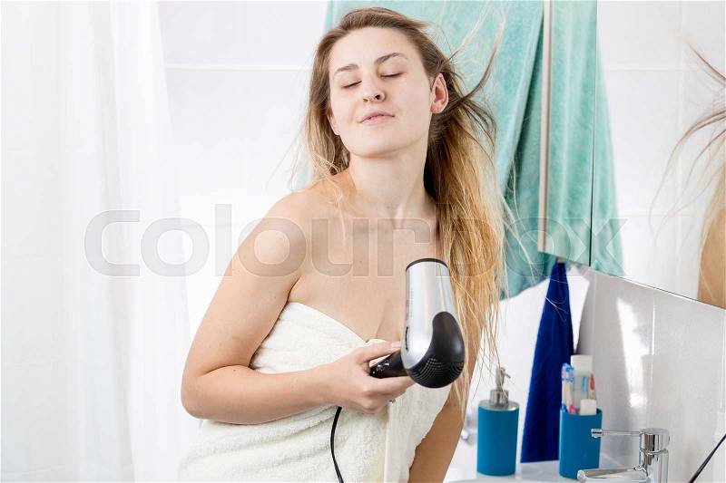 Portrait of beautiful young woman covering in bath towel drying hair with hairdryer at bathroom, stock photo