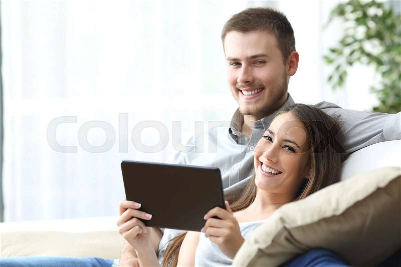 Couple holding a tablet posing sitting on a couch at home and looking at camera, stock photo