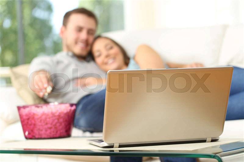 Happy couple watching tv on line in a laptop and eating popcorn lying on a comfortable sofa at home, stock photo