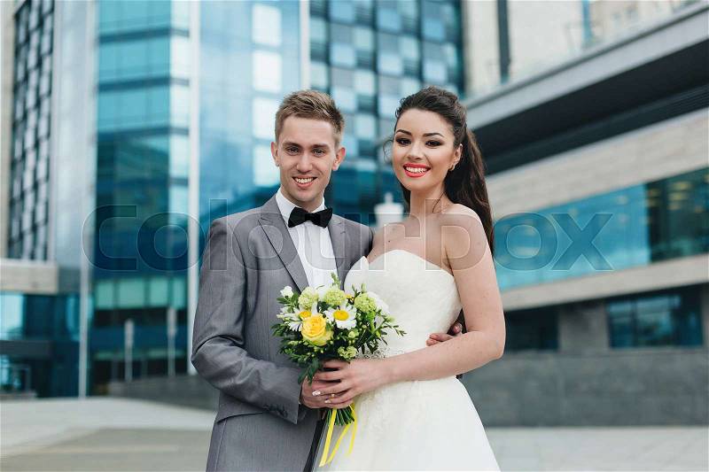 Charming bride and handsome groom smiling and holding hands in front if the high blue building, stock photo