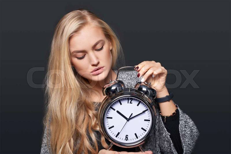Pensive woman looking at alarm clock, isolated portrait over grey background, stock photo