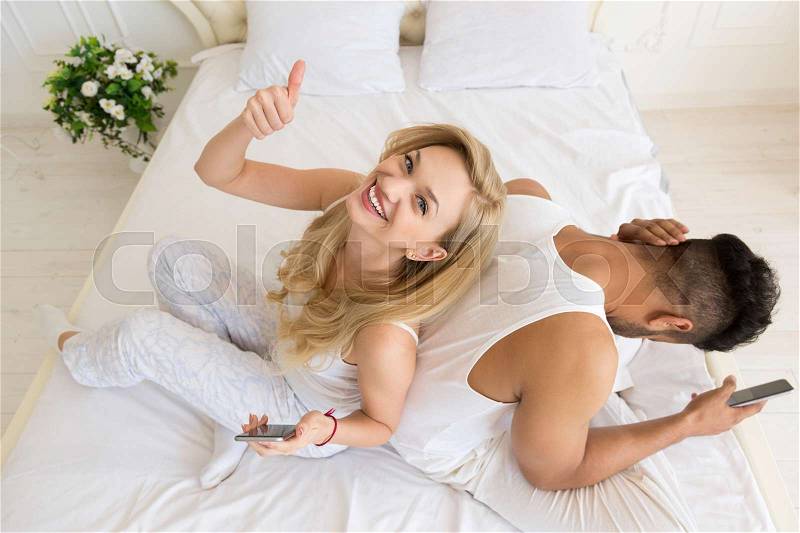 Young Couple Sitting In Bed, Happy Smile Excited Woman Hispanic Man Using Cell Smart Phone, Lovers In Bedroom Top Angle View, stock photo