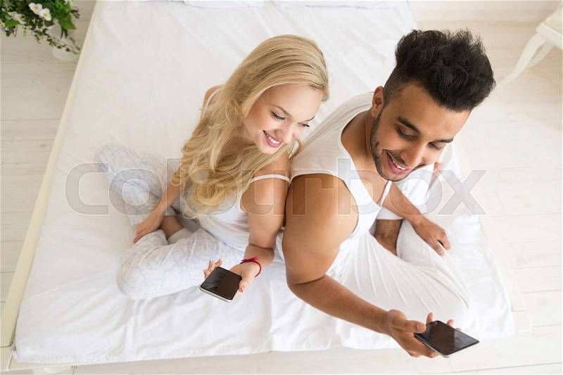 Young Couple Sitting In Bed, Happy Smile Hispanic Man And Woman Using Cell Smart Phone, Lovers In Bedroom Top Angle View, stock photo