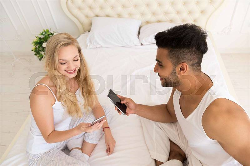 Young Couple Sitting In Bed, Happy Smile Hispanic Man And Woman Using Cell Smart Phone, Lovers In Bedroom Top Angle View, stock photo