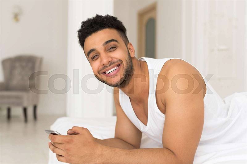 Handsome Hispanic Man Lying In Bed Using Cell Smart Phone, Young Guy Happy Smile Morning Bedroom, stock photo