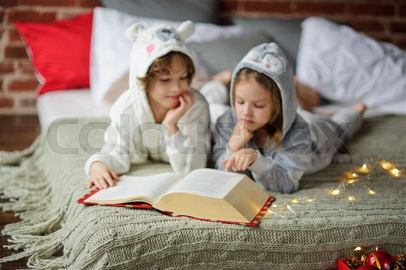 Christmas Holidays. Brother and sister lay on a bed in soft pyjamas. Bedroom is decorated by christmas garlands. Children thumb through the huge book with Christmas fairy tales. Merry Christmas, stock photo