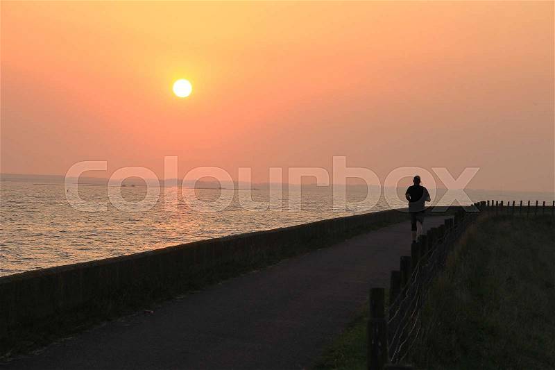 At the left side the sea and the solitary runner is training for the marathon at the dyke at the country side at sunset in the summer, stock photo