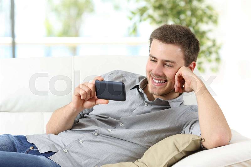 Happy guy enjoying watching entertainment content on a phone sitting on a couch at home, stock photo