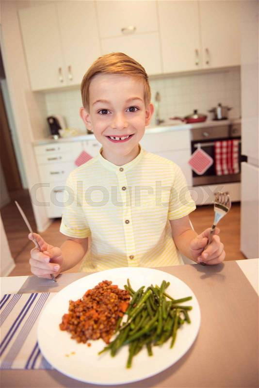 Young diligent happy boy at a table eating healthy meal using cutlery, stock photo