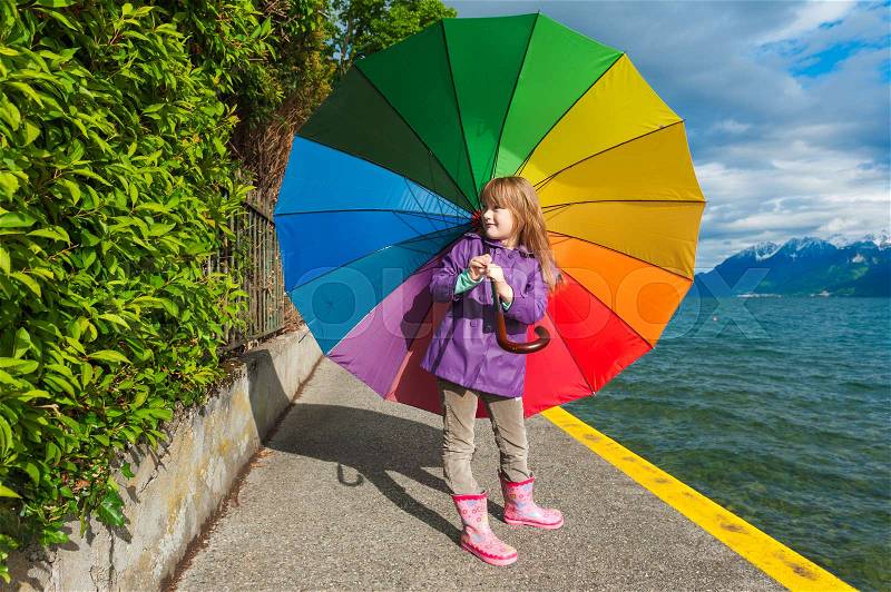 Outdoor portrait of a cute little girl with big colorful umbrella, wearing purple rain coat, boots, standing next to lake, stock photo