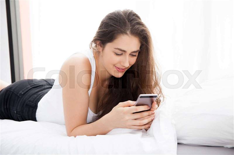 Beautiful Caucasian woman laying down on white bed using mobile phone and smile, stock photo