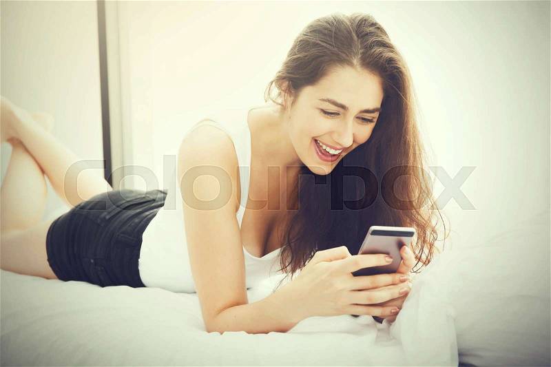 Beautiful Caucasian woman laying down on white bed using mobile phone and smile (Vintage tone), stock photo