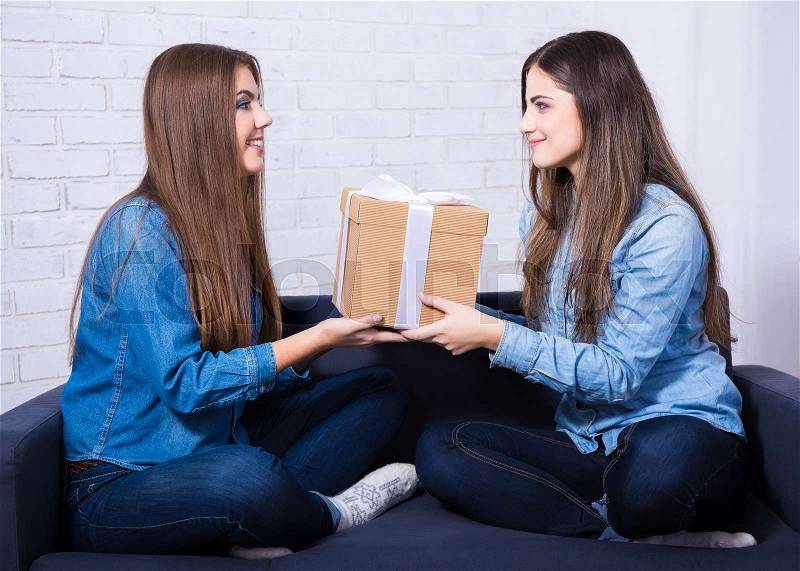 Holidays and friendship concept - two happy girls with gift box sitting on sofa at home, stock photo