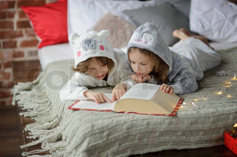 Two children, brother and sister, lie on big bed and read fairy tales. Children dressed in fluffy pajamas, like little cute animals. Before them the huge book. Children are keen on reading, stock photo
