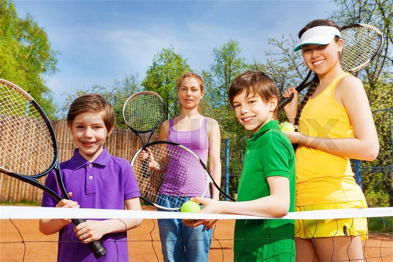 Happy sportive family of four people, standing near tennis net on the clay court with rackets in summer, stock photo
