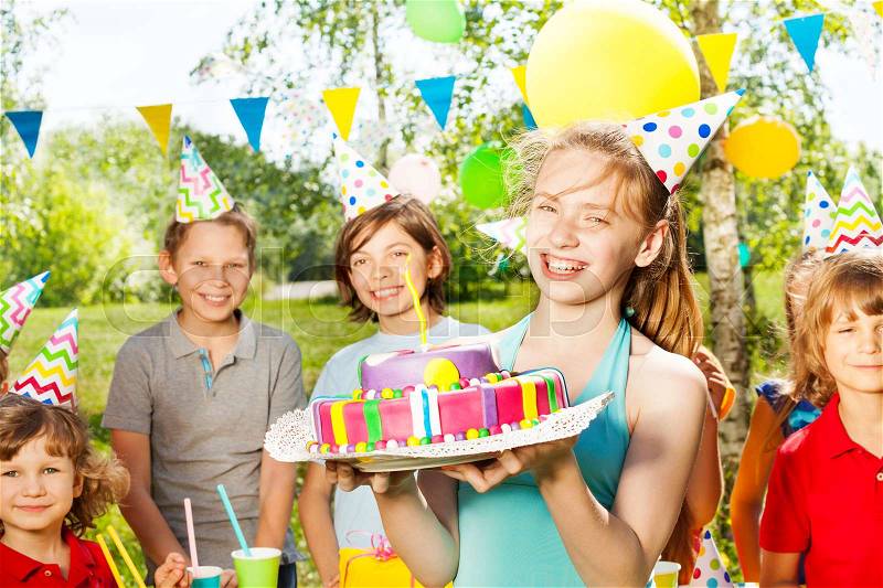 Laughing young girl in party hat holding birthday cake, standing among her friends at the outdoor party, stock photo