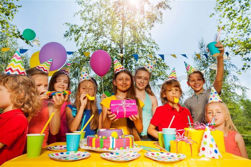 Funny kids in party hats cheering birthday girl, blowing the whistles at summer park, stock photo