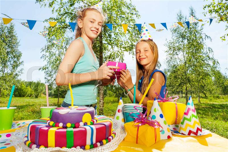 Happy young girl giving birthday gift to her friend, standing next to the festive table, at the outdoor birthday party, stock photo