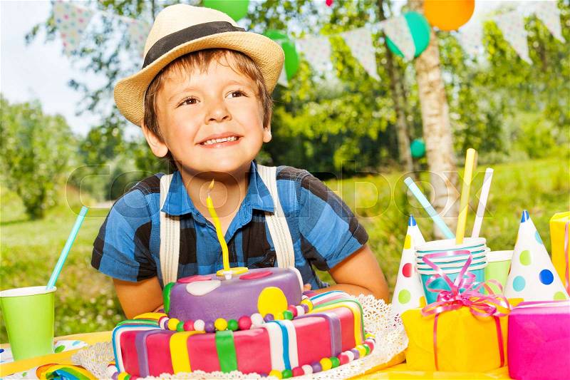 Portrait of happy smiling kid boy making a wish, sitting next to the birthday cake at the outdoor party, stock photo