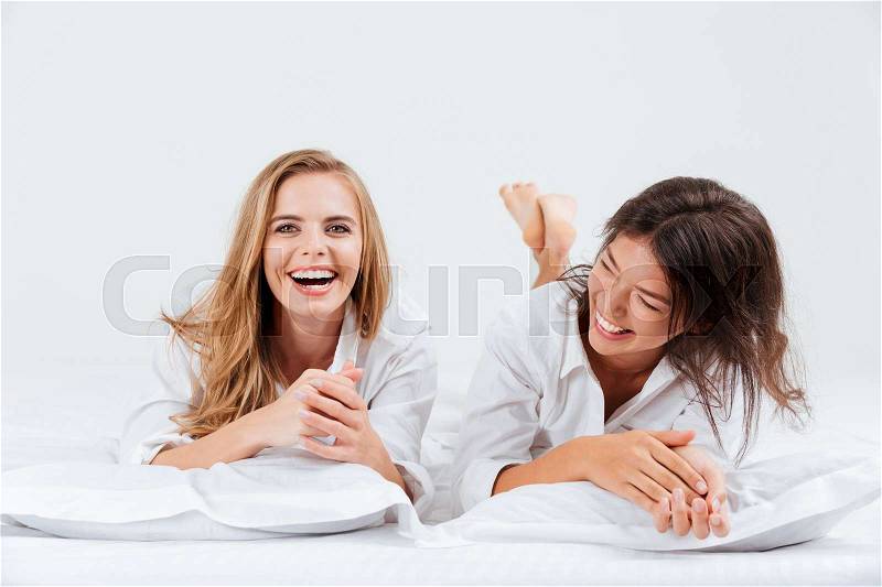 Close up portrait of two laughing beautiful woman lying together on bed with white sheets, stock photo