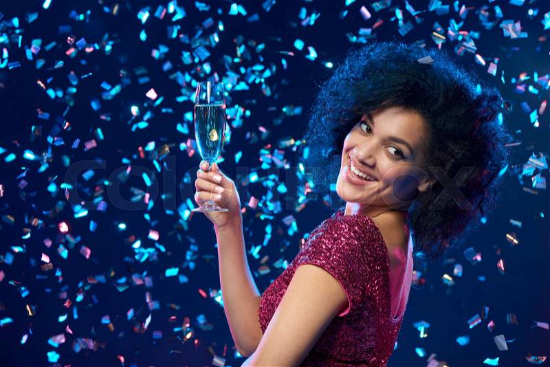 Party, drinks, holidays and celebration concept. Closeup of happy mixed race woman in sequined dress with glass of sparkling wine over colorful background with confetti looking to the side at blank copy space, stock photo