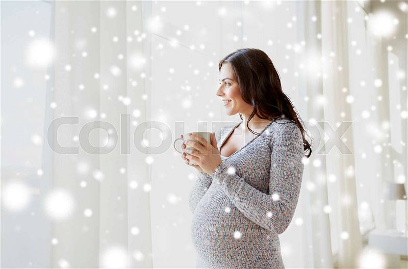 Pregnancy, drinks, winter, people and expectation concept - happy pregnant woman with cup drinking tea looking through window at home over snow, stock photo