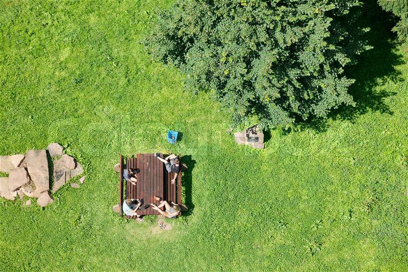 Friends resting in the nature in a green field after walking or hiking gathered around at a table sitting and talking. Outdoor scene in summer countryside, view from above, stock photo