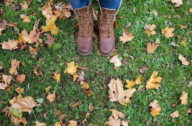 Season, footwear and people concept - female feet in boots with autumn leaves on grass, stock photo