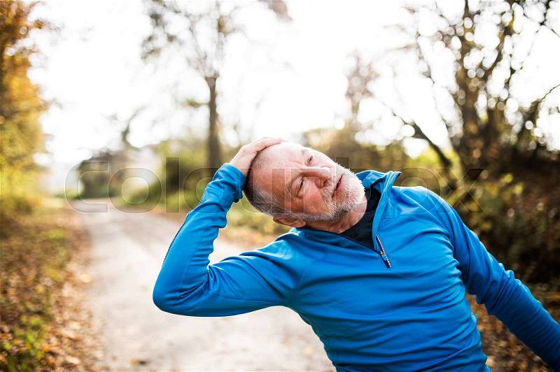 Senior runner in sunny autumn nature doing stretching. Man with gray beard and mustache wearing blue sweatshirt exercising, stock photo