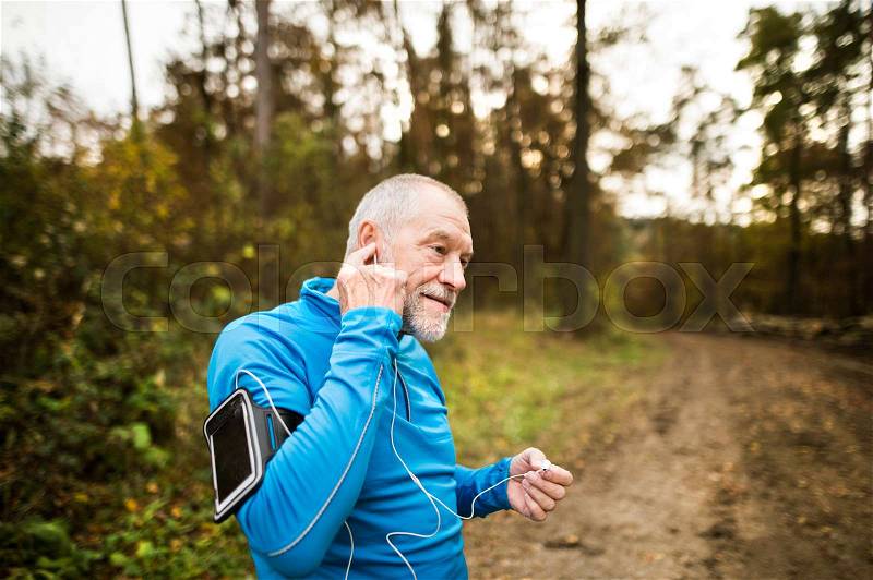 Senior runner in nature. Man with smart phone, putting earphones into his ears. Listening music or using a fitness app. Using phone app for tracking weight loss progress, running goal or summary of his run, stock photo