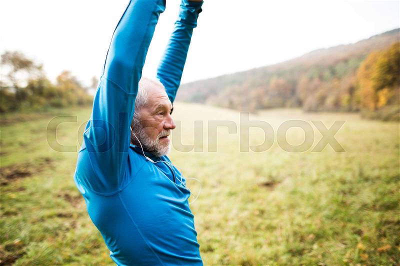 Senior runner in sunny autumn nature with earphones, listening music, doing stretching, stock photo