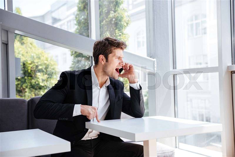 Mad angry young businessman sitting in cafe and shouting in cell phone, stock photo