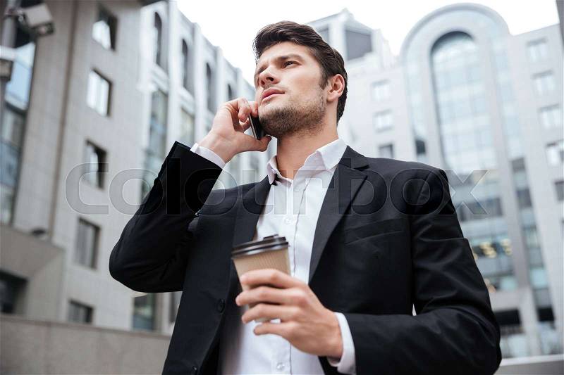 Confident young businessman with take away coffee talking on cell phone near business center, stock photo
