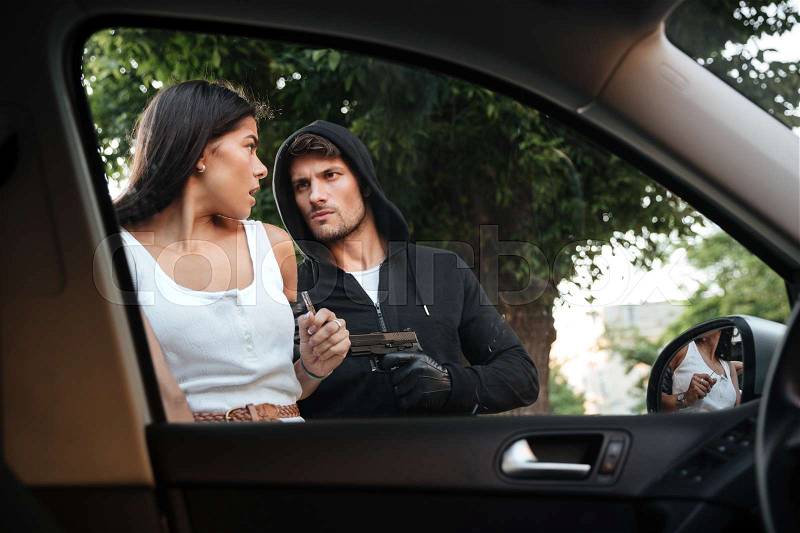 Dangerous criminal man with gun stealing car of scared young woman on outdoor parking, stock photo