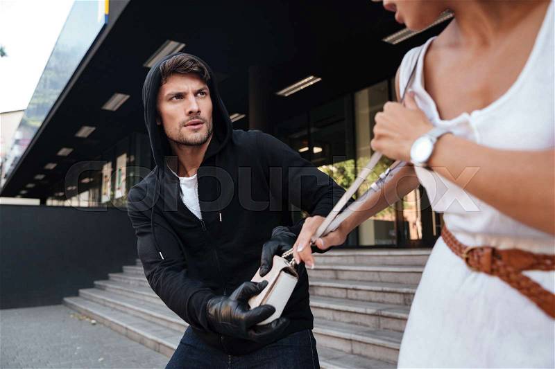 Criminal man in hoodie stealing woman bag on the street, stock photo