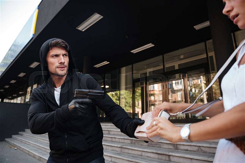 Man robber in hoodie stealing woman bag and threatening with gun on the street, stock photo