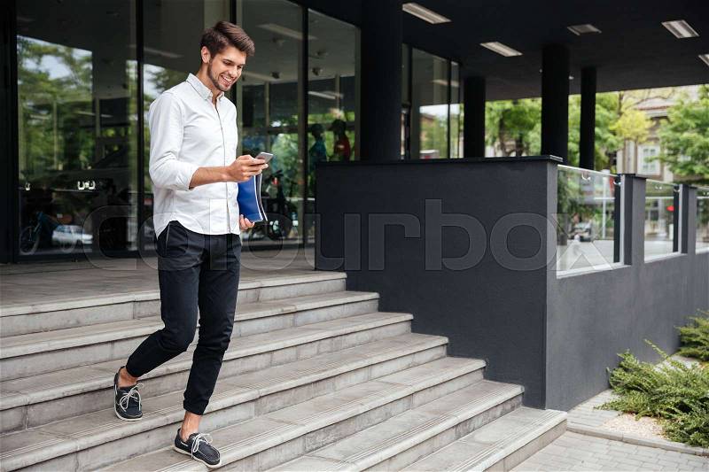 Smiling attractive young businessman walking and using mobile phone near business center, stock photo