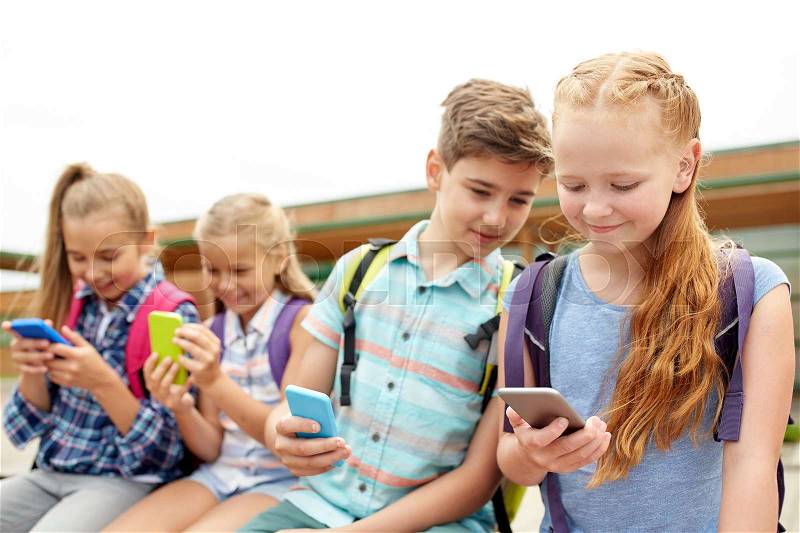 Primary education, friendship, childhood, technology and people concept - group of happy elementary school students with smartphones and backpacks sitting on bench outdoors, stock photo