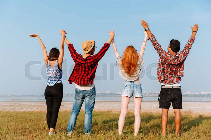 Back view of group of young friends standing and holding raised hands outdoors, stock photo