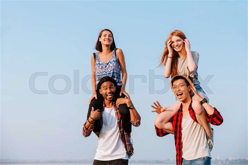 Two cheerful young women sitting on shoulders of their boyfriends outdoors, stock photo