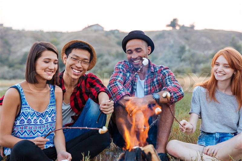 Group of happy young people sitting and roasting marshmallows on campfire outdoors, stock photo