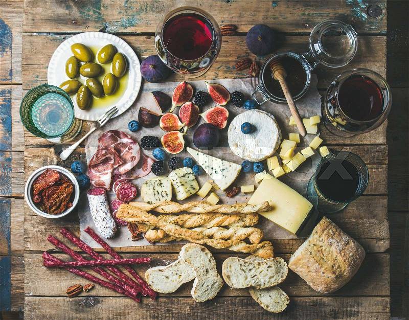 Wine and snack set with various wines in glasses, meat variety, bread, sun-dried tomatoes, honey, green olives, figs, nuts and berries on wax paper over rustic wooden table background, top view, stock photo
